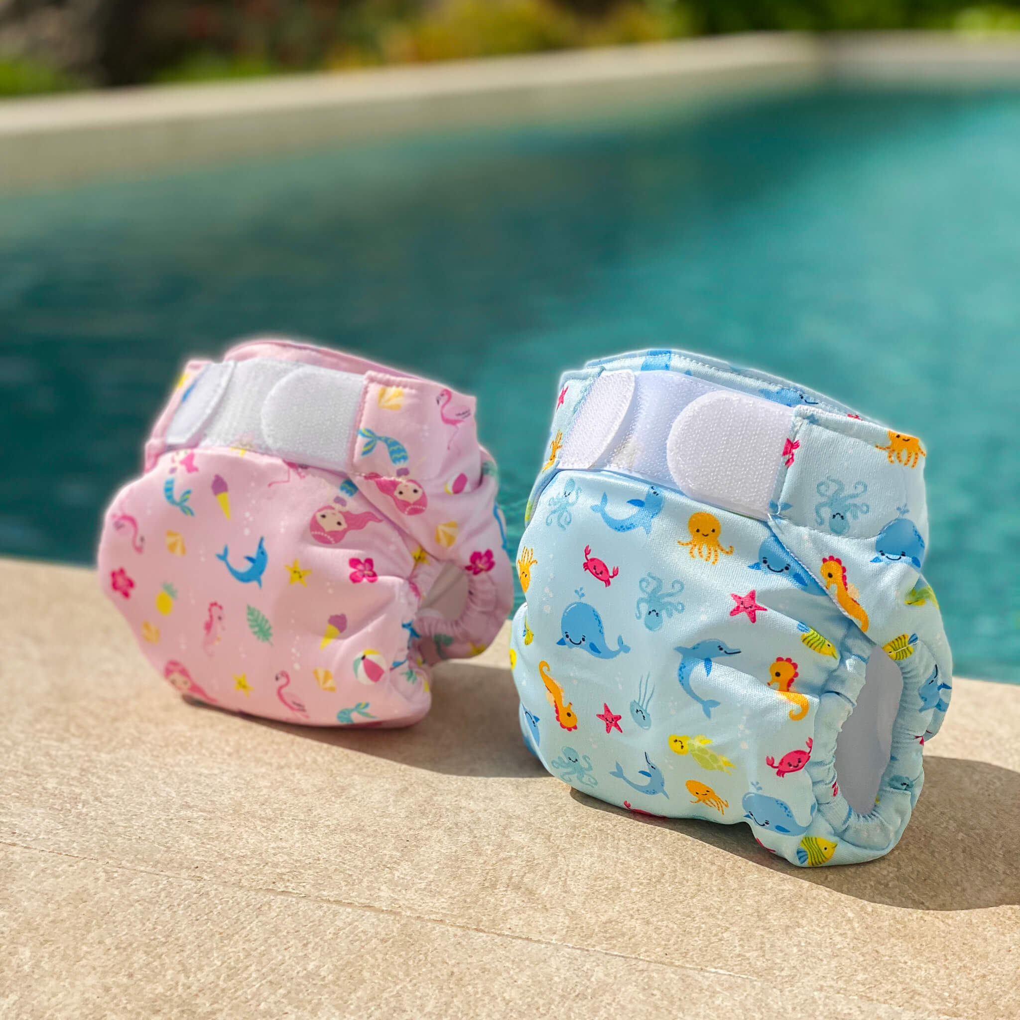Pannolini-costume lavabili Swim Nappy rosa e blu davanti a una piscina.